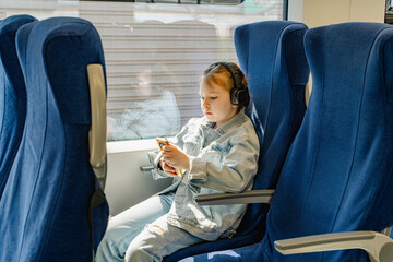 Little girl traveling on train in headphones looking at mobile phone. A little traveler on a train...