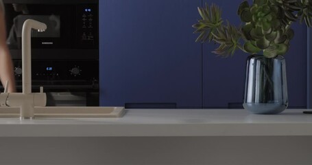 Modern blue kitchen interior design with build-in cabinet stone island sink faucet. Young woman walks through kitchen island touches stone countertop with hand