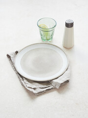 Table set, empty white and brown plate on napkin, salt shaker and light green glass with lemon on a light-colored background.