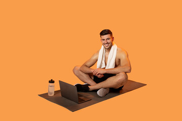 A fit man with a towel around his neck is seated cross-legged on a yoga mat, engaged in an online...