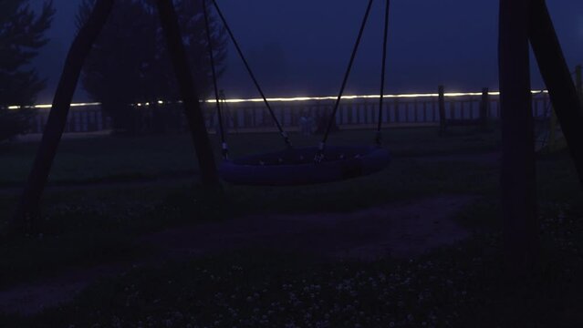 Empty swing on children's playground swinging at foggy night and two girls sitting on the grass on the background. Horrible poltergeist and ghost concept	