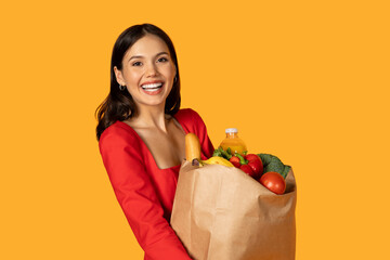Happy young woman is standing outdoors, holding a paper bag filled with groceries. She appears to...