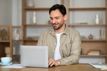 A man is seated at a table, focused on his laptop screen. Beside him, there is a steaming cup of...