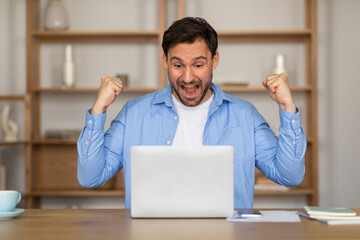 A man is energetically celebrating in front of a table while holding a laptop. He appears to be...