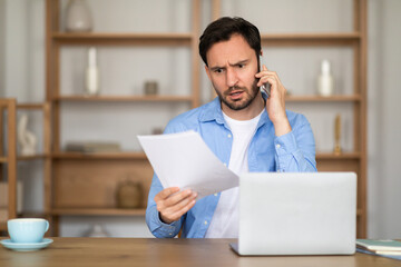 A man appears engaged in a serious conversation on his cellphone, simultaneously examining a document with a furrowed brow. The scene takes place in a well-lit home office