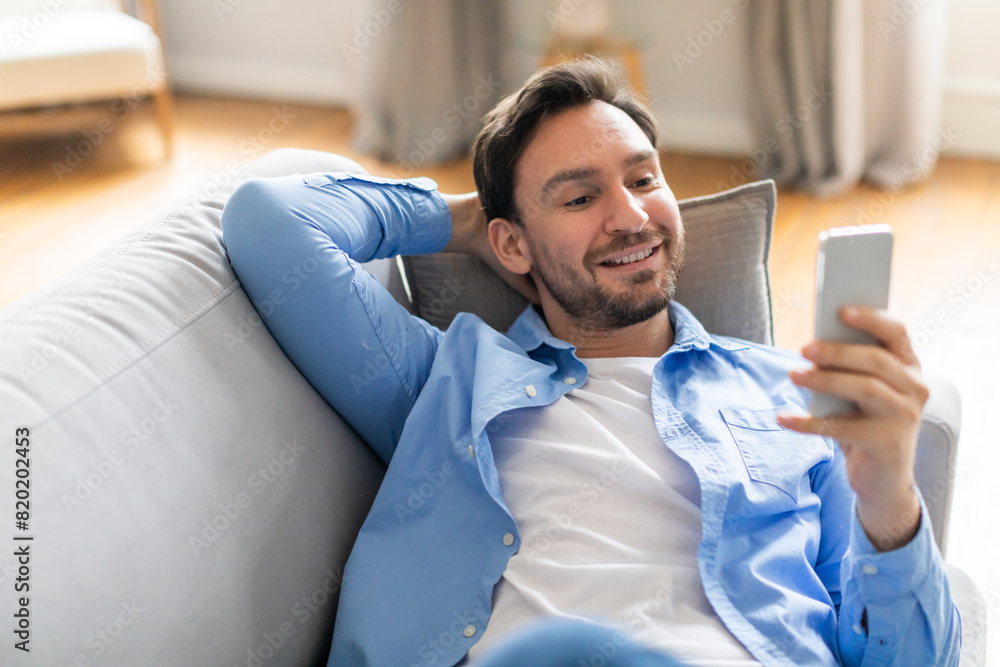 Poster A man is seated on a couch, engrossed in his phone screen. He appears focused as he scrolls and interacts with the device.