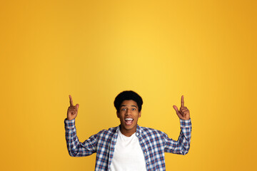 Cheerful Teen Guy Pointing Upwards and Looking at Camera on Orange Studio Background