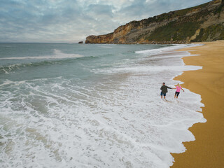 A young couple walks on the beach of Nazaré during the waves on a spring day, photo from a drone.