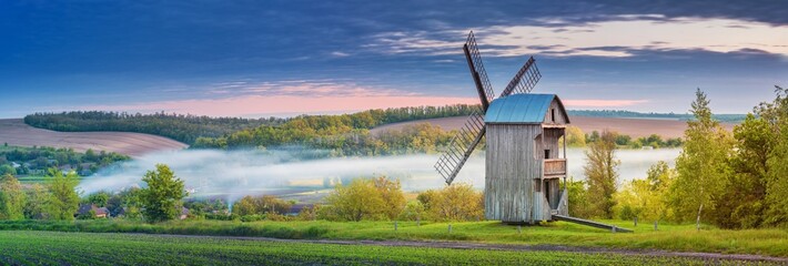 Beautiful panoramic landscape of spring morning in farmland with old wooden windmill and fog in...
