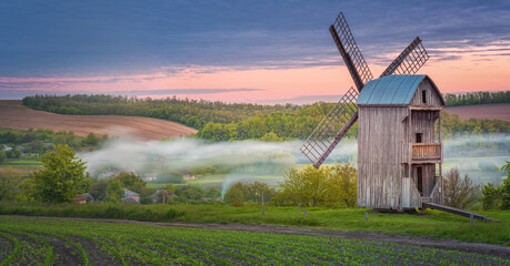 farm landscape with old wooden windmill and fog in village in morning twilight