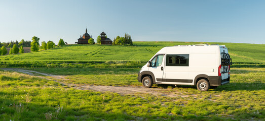 panoramic view to white camper van in country location with old wooden church in spring day