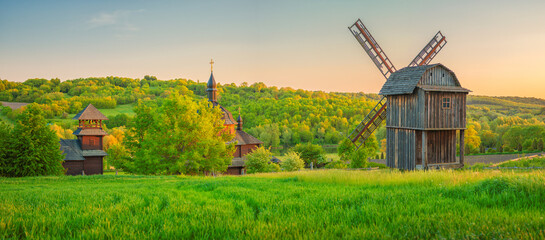 panorama landscape of farmland with old wooden windmill and church in warm light of sunset