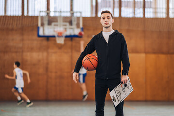 A young basketball coach standing at court with ball and clipboard.