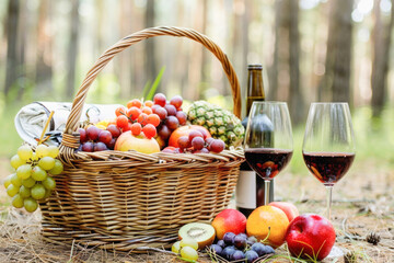 A picnic basket is brimming with fresh fruit and accompanied by two glasses of wine on the grass