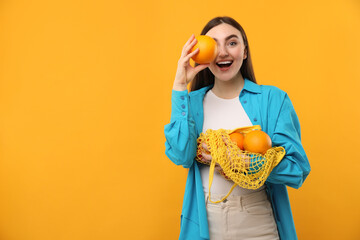 Woman holding string bag of fresh oranges and covering eye with fruit on orange background, space...