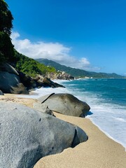 photo d'une plage paradisiaque avec une eau turquoise et des traces de pas sur le sable. Des gros rochers sur le sable fin, les vagues arrivent sur les rochers. La végétation est luxuriante, Tayrona 