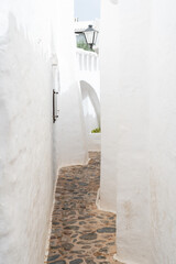 A picturesque view of an empty street in Binibeca, Menorca. The whitewashed buildings and cobblestone pathway showcase the charming Mediterranean architecture of this tranquil village