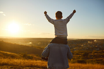 Little cute child boy sitting on his fathers shoulders looking into the distance enjoying landscape,sunset and beautiful nature outdoors. Son walking with dad in the field. Fathers day concept