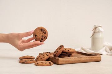 Female hand with wooden board of sweet cookies with chocolate chips and bottle of milk on white...