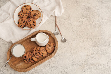 Wooden tray and plate of sweet cookies with chocolate chips and jug of milk on white background