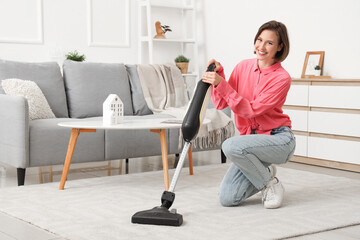 Happy young woman cleaning carpet with handheld vacuum cleaner in living room