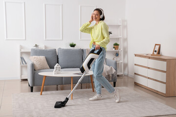 Happy young woman in headphones cleaning carpet with handheld vacuum cleaner in living room