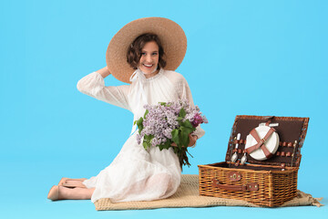 Beautiful young woman with bouquet of blooming lilac flowers and wicker picnic basket sitting on...