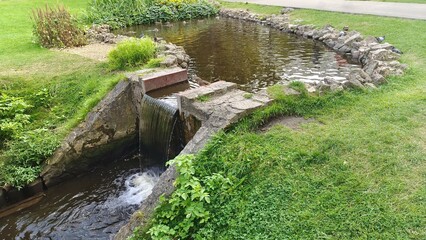 On a grassy lawn in the city park, a stone and concrete cascade of ponds with waterfalls sits next to a walking path. Pigeons walk on the grass and ducks swim in the water. Warm summer weather
