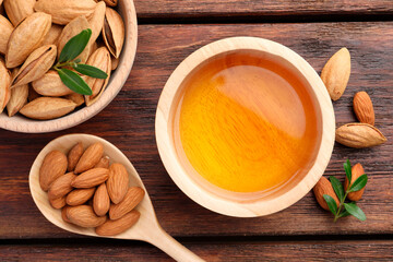 Almond oil in bowl, nuts, spoon and leaves on wooden table, flat lay