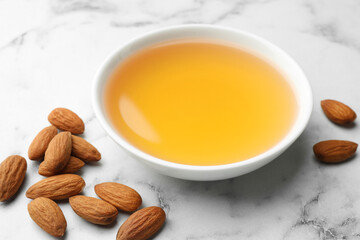 Almond oil in bowl and nuts on white marble table, closeup