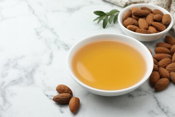 Almond oil in bowl and nuts on white marble table, closeup. Space for text
