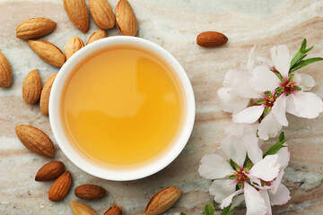 Almond oil in bowl, branch with flowers and nuts on color marble table, flat lay