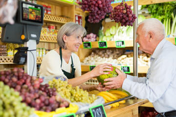 Woman supermarket employee hands watermelon to an elderly man after paying for purchase at the checkout