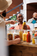 Black woman looks curiously and listens to bearded man while holding glass bottle filled with an organic yellow liquid in an eco friendly store. Female client interested in purchasing bio pasta sauce.