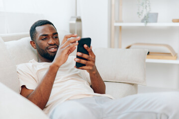 Happy African American man holding a black smartphone, sitting on a modern white sofa in his...