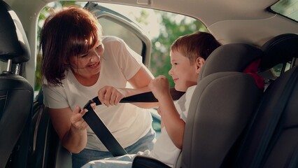 Caring mother placing son to car childish armchair and fasten seat belt for protection in vehicle. Family road trip compliance with safety rules. Woman parent and boy child at automobile safe control
