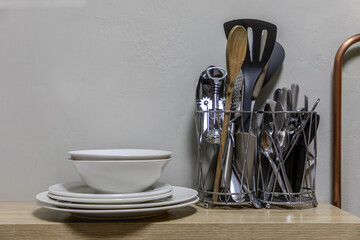 Selection of kitchen utensils on a shelf with plates and bowls stacked next to it against a...