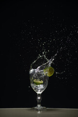 a slice of lemon falling into a glass cup with clear water causing splashes. Isolated on dark background.