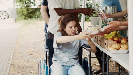 Caucasian wheelchair-bound woman receiving free food from charity workers at outdoor non-profit...