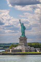 Fototapeta premium New York, NY, USA - August 1, 2023: Statue of Liberty isolated on portrait under blue cloudscape. Green island and plenty of visitors add colors
