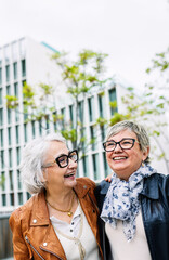 Two senior adult women having fun together at city street. Friendly female friends laughing while hugging each other walking outdoors. Mature friendship and retired people concept.