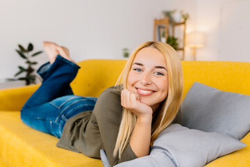 Happy young woman relaxing on her couch at home in the living room. Smiling portrait of blonde female looking at camera lying on sofa.