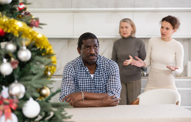 Upset middle-aged man sitting at table while two women quarreling to him standing behind in the...