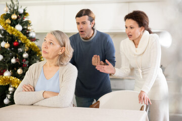 Couple actively scold mother who listens and thinks about it for сhristmas, sitting at table in kitchen near christmas tree