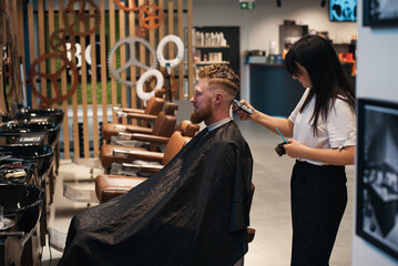 Red-haired bearded man sitting in barbershop and female barber making hairdo to guest