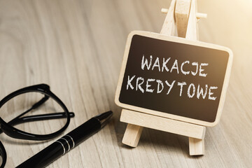 Black writing board on a wooden frame with the inscription "wakacje kredytowe", next to black glasses and a pen (selective focus) translation: credit holidays