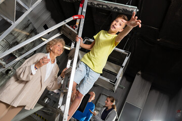 Young boy standing on stepladder and reaching his hand in escape room. His grandmother standing...