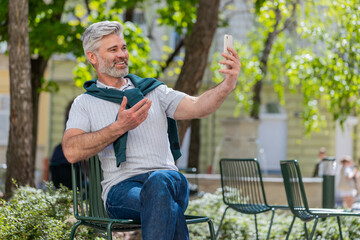 Obraz premium Bearded mature man blogger using smartphone, communicating video call online with subscribers, recording stories for social media vlog outdoors. Guy sitting on chair in urban sunshine city street.