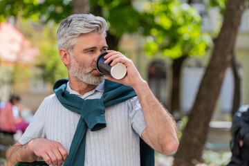 Happy Caucasian mature man enjoying morning coffee hot drink sitting on cafe chair. Relaxing, taking a break. Bearded guy on urban city center street, drinking coffee to go. Town lifestyles outside.