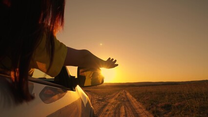 Beautiful girl with sits in front seat of car, her hand out window, catching glare of setting sun. Free young woman travels by car, extends her hand to sun from car window. Vacation. Woman driver sun